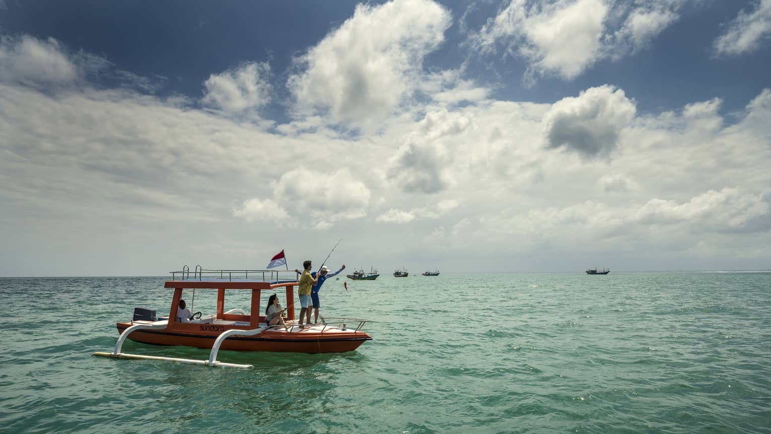 On the open water, three people fish from the hull of an outrigger-style boat; four other boats float in the distance.