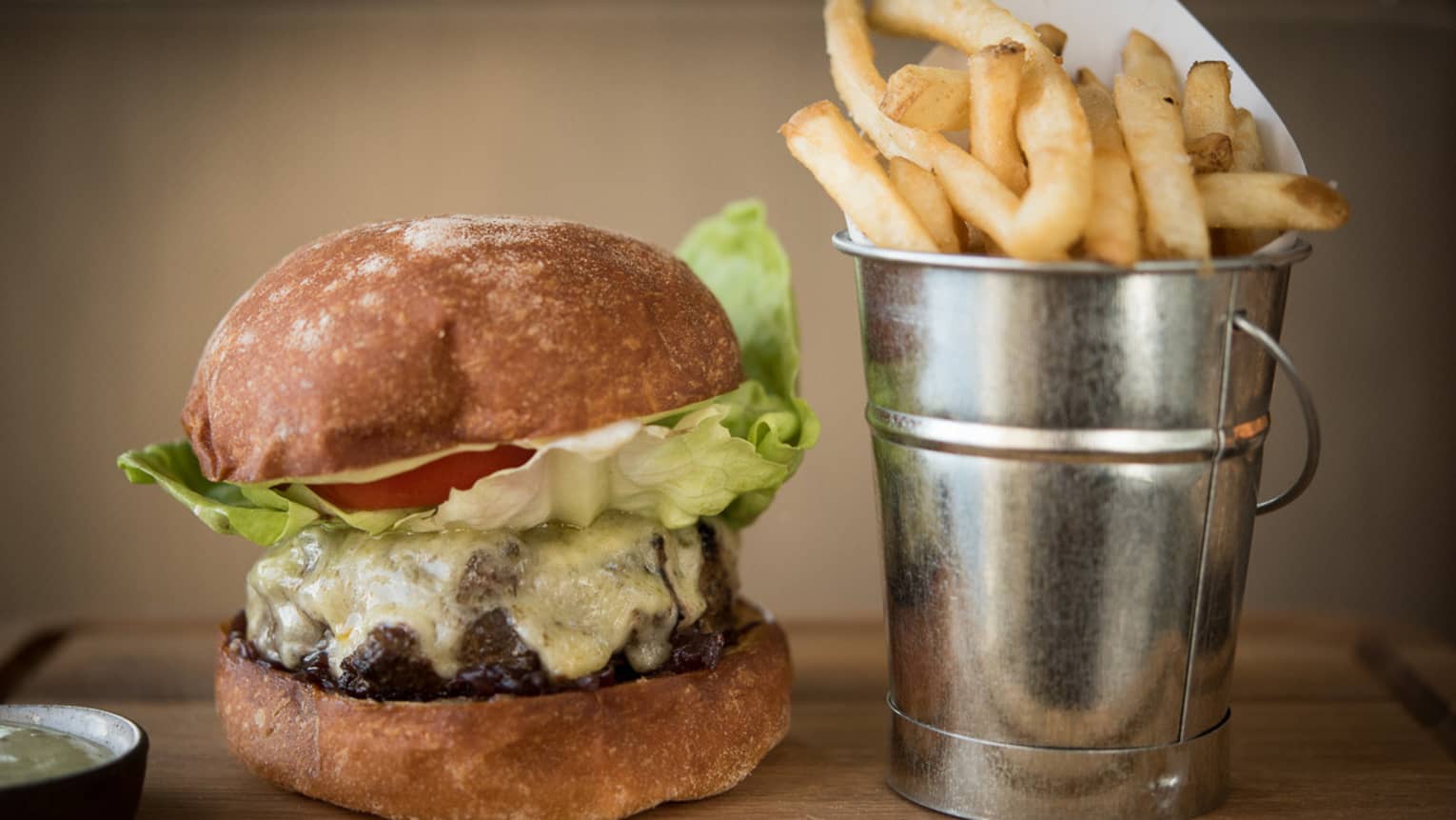 Thick beef burger with melted cheese on artisan bun beside silver tin with French fries
