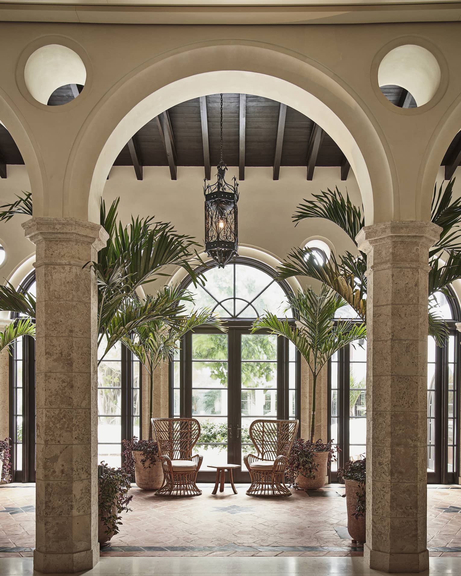 An archway looking at two wood chairs by a large window and small table, there are palm trees around the chairs.
