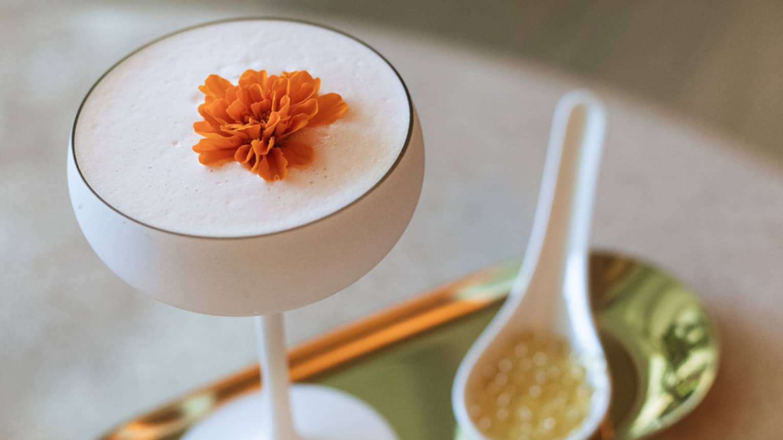 White opaque coupe glass filled with a cocktail and topped with an orange flower