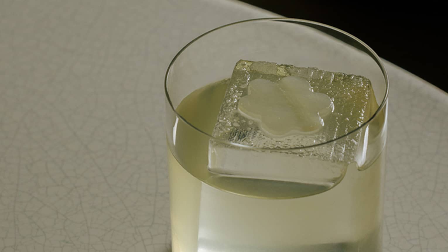 Light-coloured cocktail in a rocks glass with one large ice cube in the glass