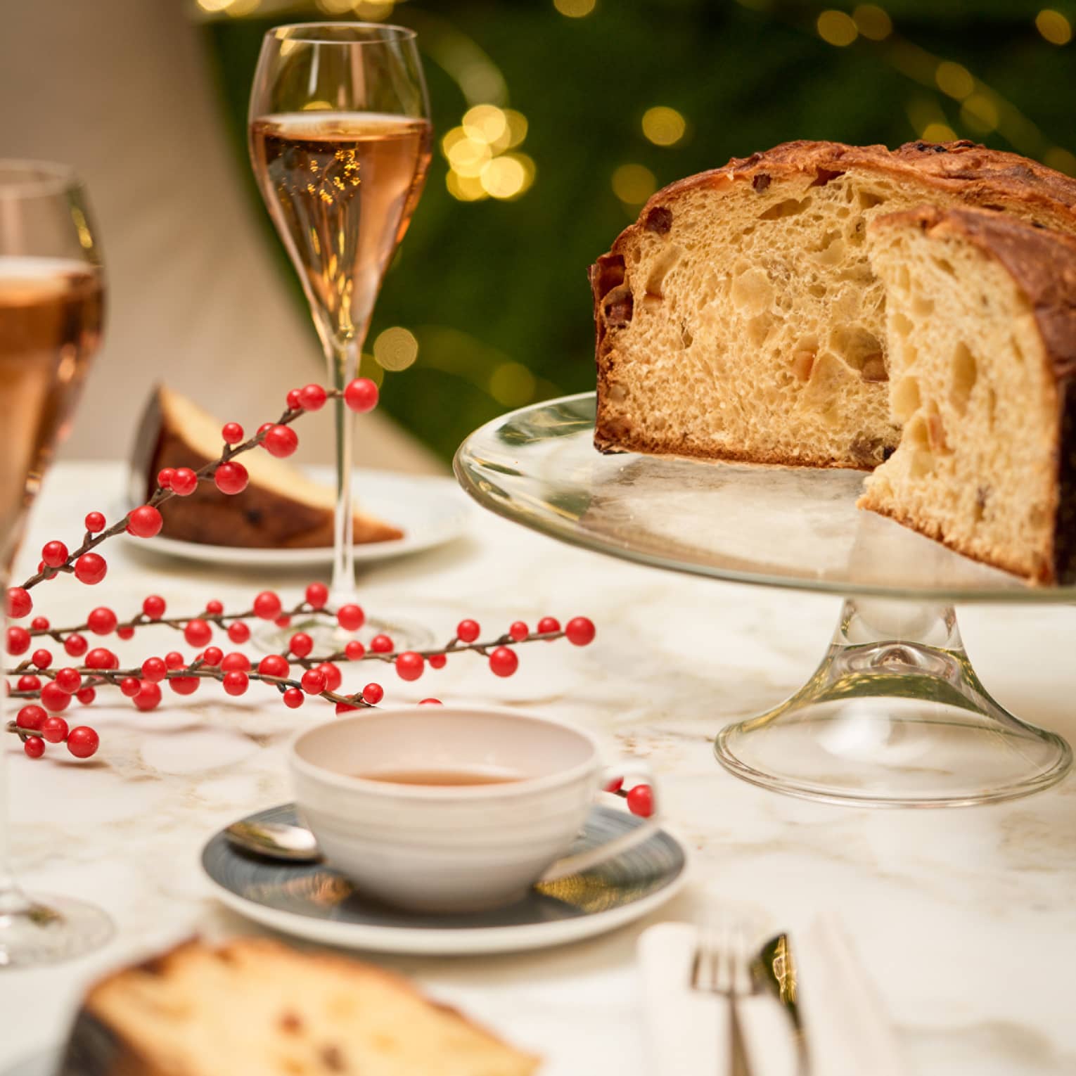 Loaf of panetonne sits on a crystal cake stand with two slices plated on the table next to it, two glasses of pink-hued wine in champagne flutes and a cup of tea
