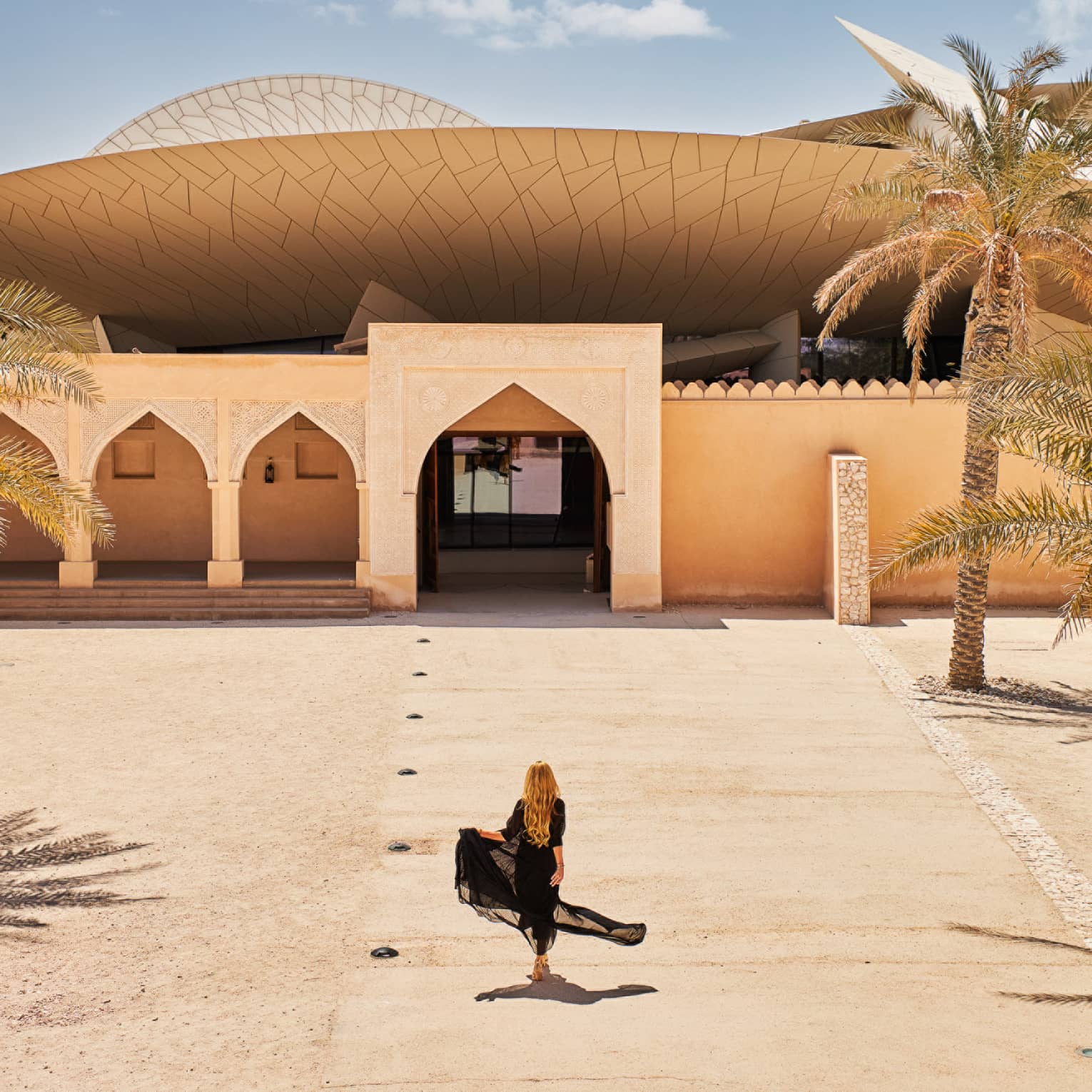 Person wearing long black flowing dress walks along a sandy walkway leading up to an Arabesque-style building
