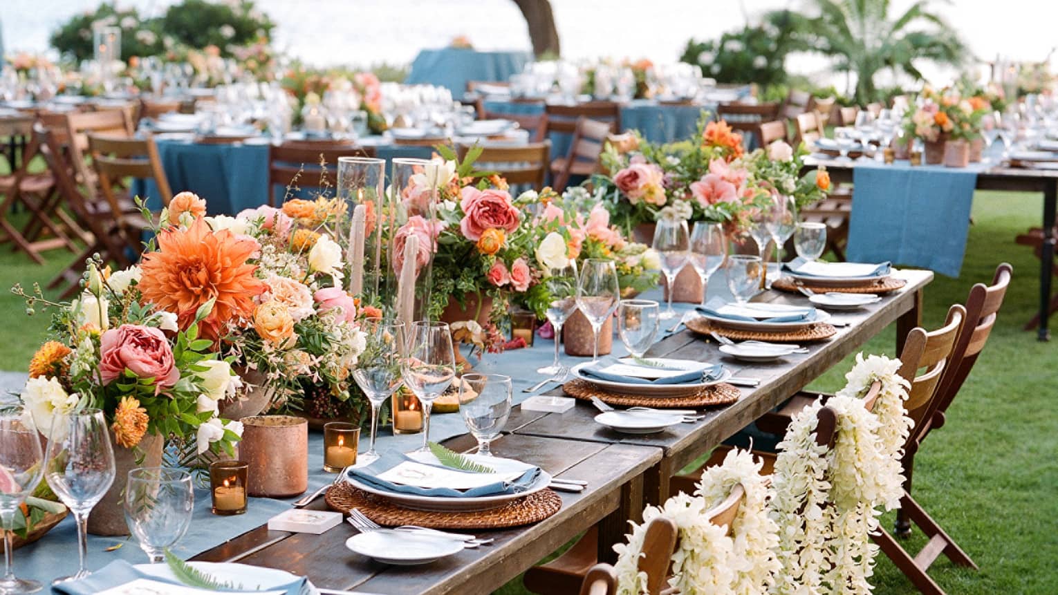 Close-up of outdoor banquet dining table with flower arrangements, white floral leis draped over chairs