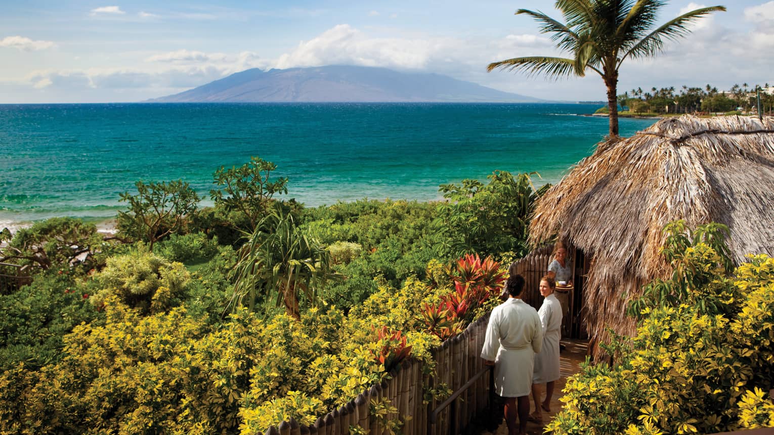 Couple in white bathrobes follow spa attendant into thatched-roof hut on path looking out at ocean
