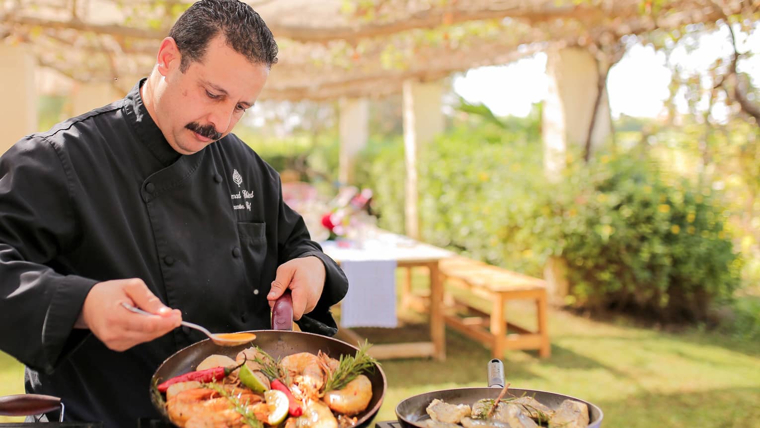 Chef Emad Ebeid pours sauce over seafood dish in pan at outdoor kitchen