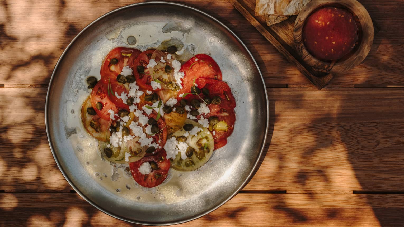 A round plate holds a colorful mixture of tomato slices, capers and white cheese crumbles. Next to it is a side of bread and a small bowl holding a condiment.