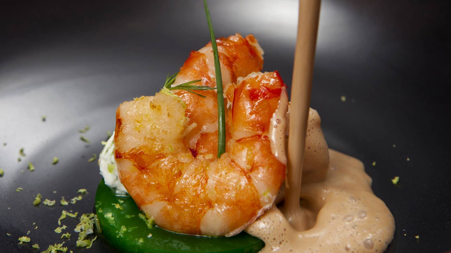 Close-up shot of sauce being pouring into a shallow bowl containing whole prawns with chive and dill garnishes