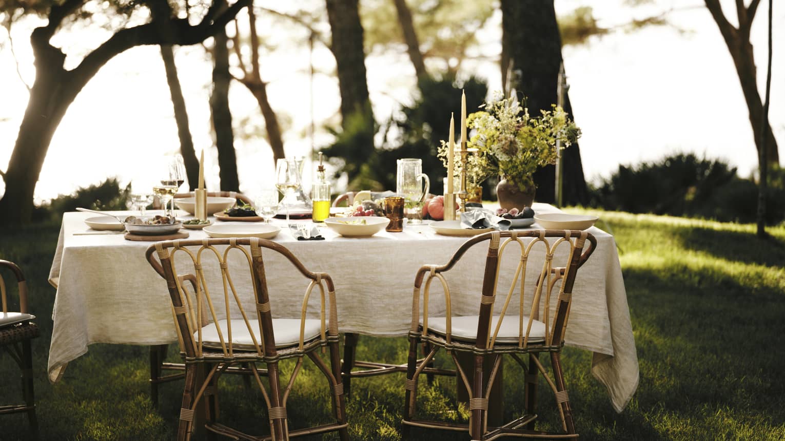 A linen tablecloth adorns an outdoor table set for five with wine, fresh seafood and fruit in an ocean-view forest setting.