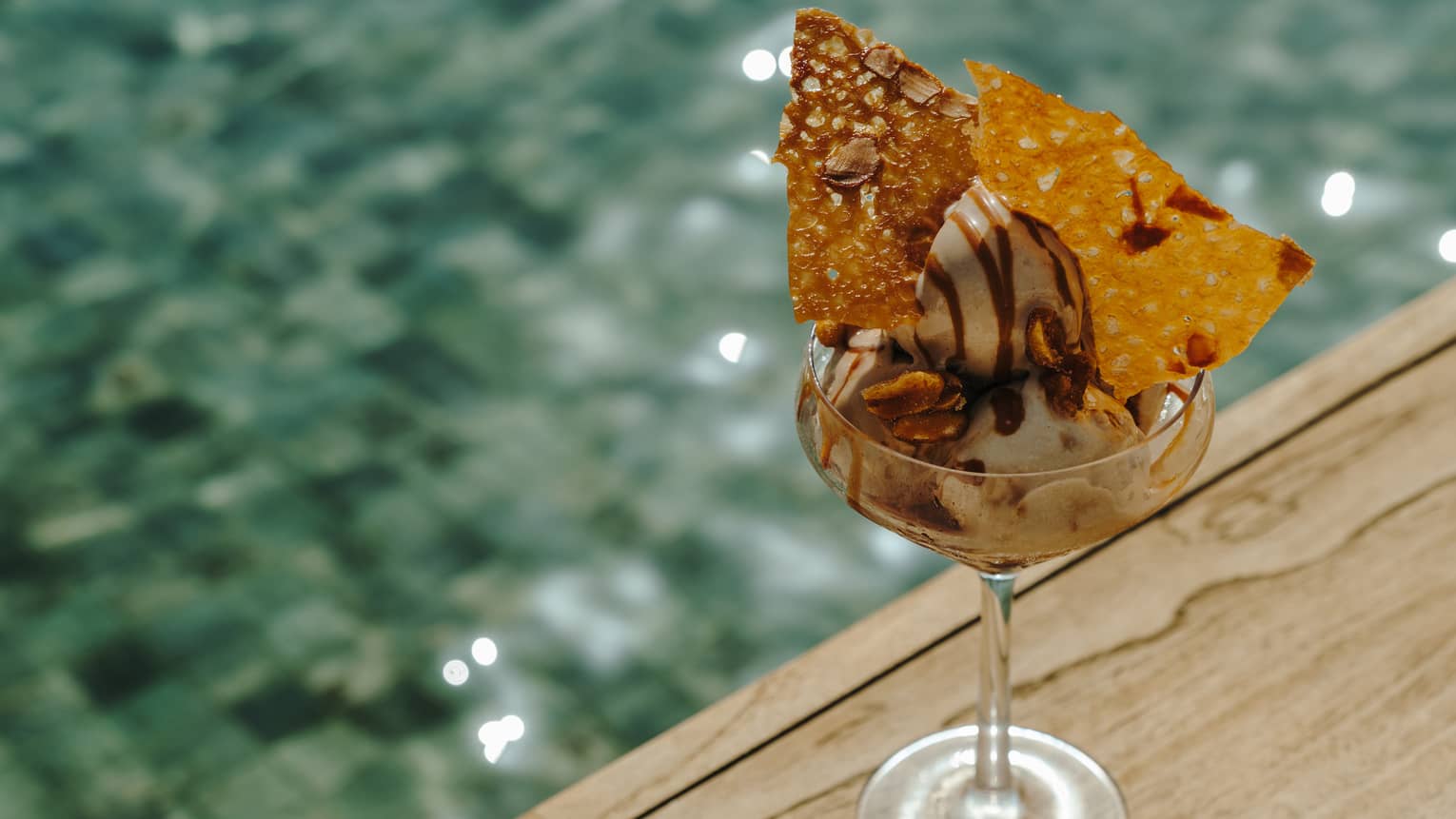 A stemmed glass sitting near the corner of a wood table holds ice cream with garnishes.