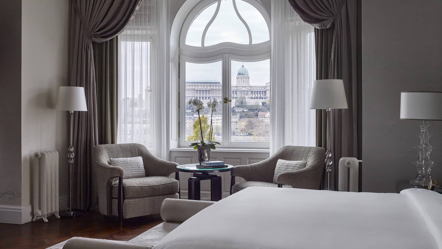 Buda Castle Presidential Suite is furnished with a two sitting chairs next to a curved window, lamps, a large bed and gray curtains. 