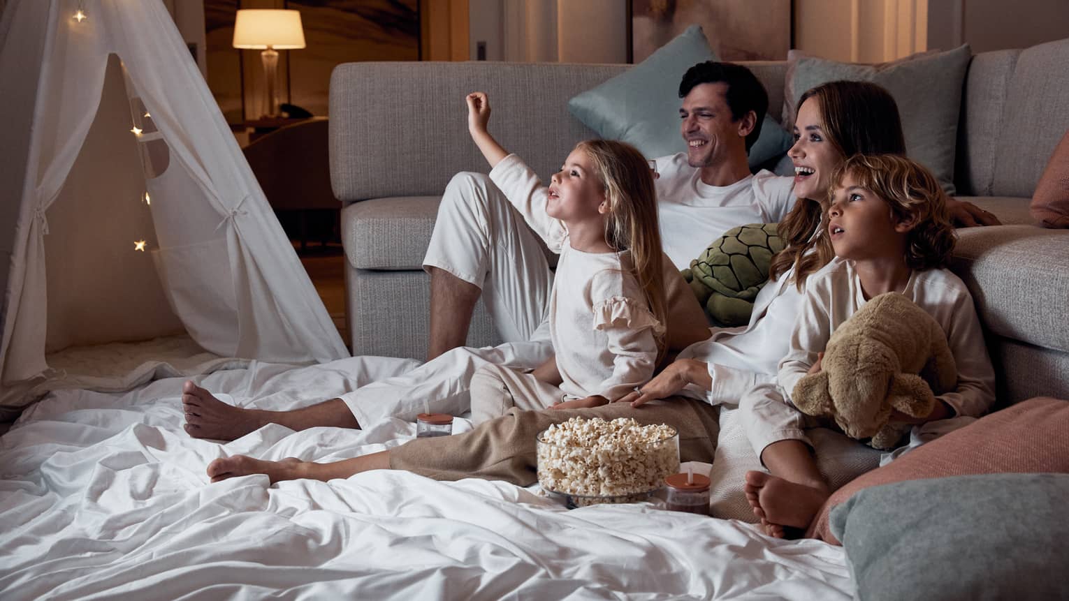 A family relaxing on blankets beside a sofa; the children hold stuffed toys and there's a bowl of popcorn between them.
