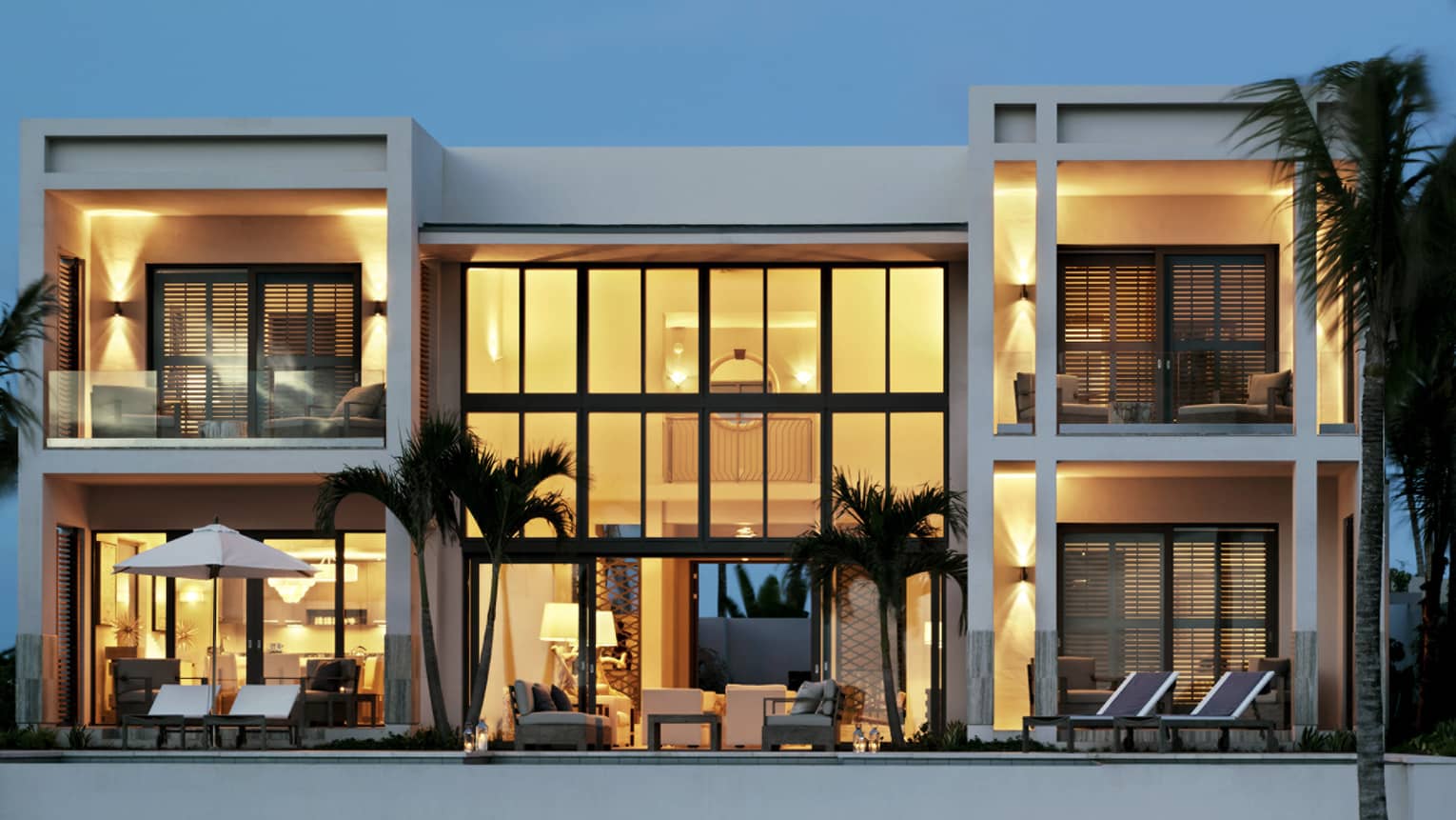 Exterior view of five-bedroom, white beachfront villa at dusk, two storeys with floor-to-ceiling illuminated windows