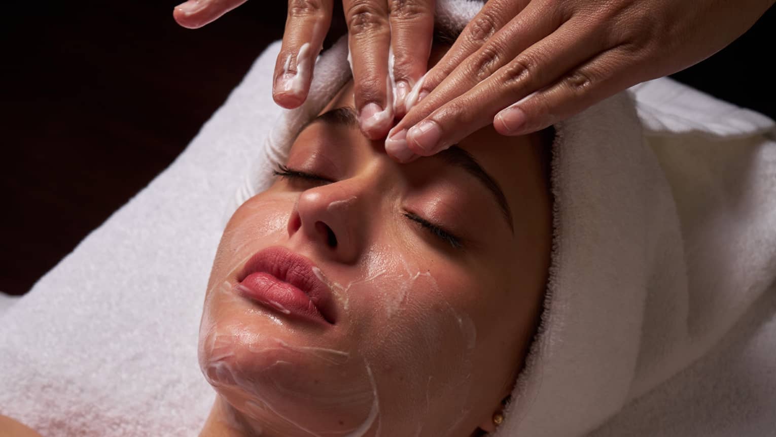 Person lies on massage table covered in a white cloth from shoulders down with hair wrapped in white towel while a pair of hands massages their forehead