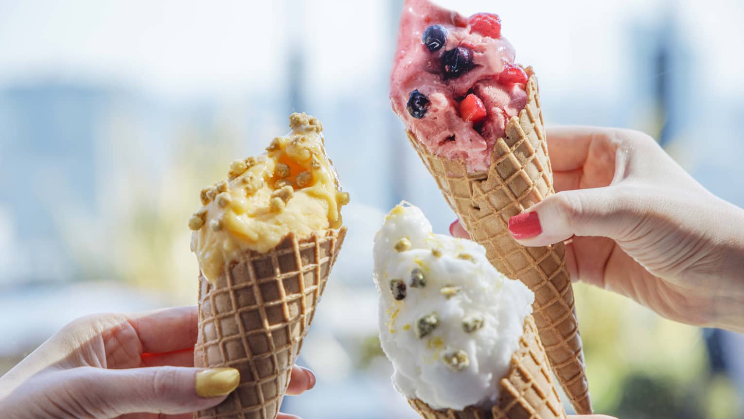 Three hands each hold a cone of ice cream in assorted flavours and colours