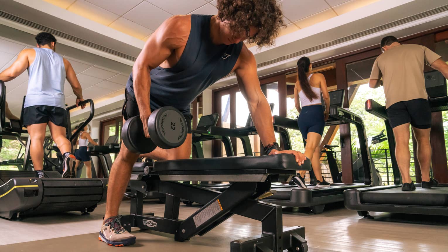 Person stands with one knee on a bench as they lift a dumbell with one arm, as three others exercise behind them on treadmills