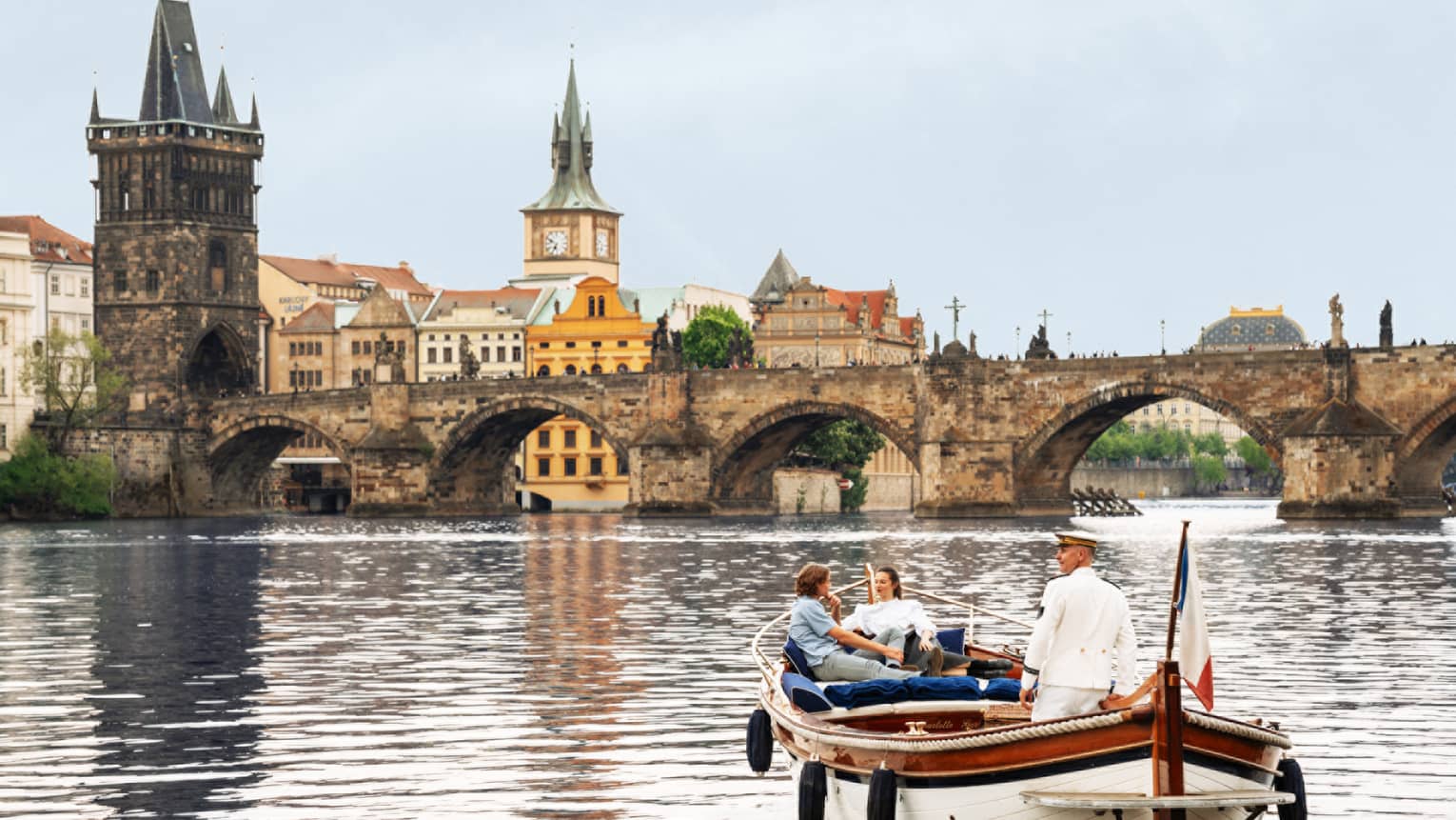 Couple with captain in wooden boat on Vltava River with bridge and buildings in backdrop