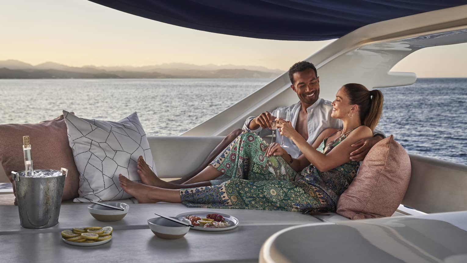 Aboard a boat in open air under a sunshade, a cuddling couple clink wine glasses amid cushions, dishes and an ice bucket.