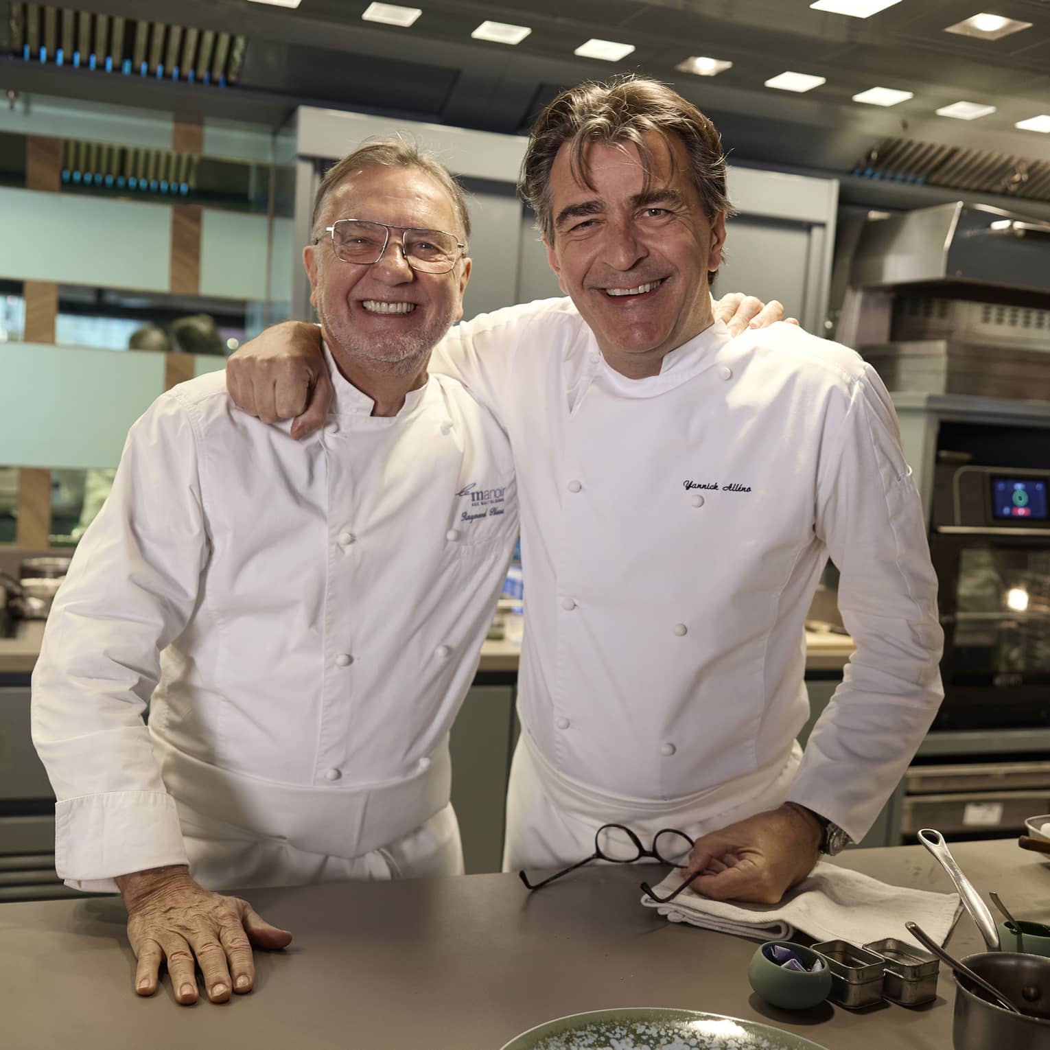 Two chefs in white uniforms, standing in front of a counter in a restaurant kitchen