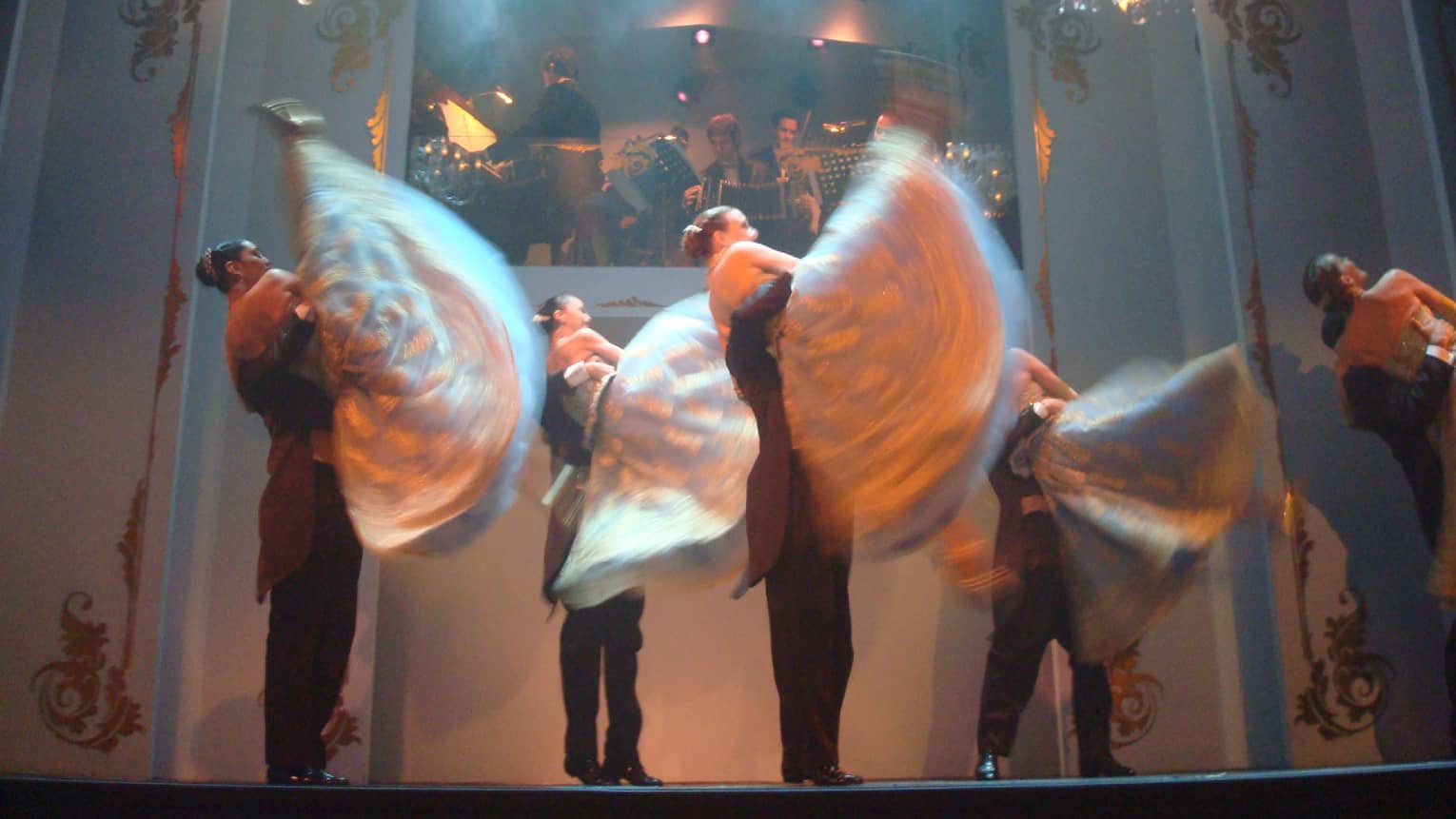 Tango dancers twirl on stage under small chandeliers, lights 
