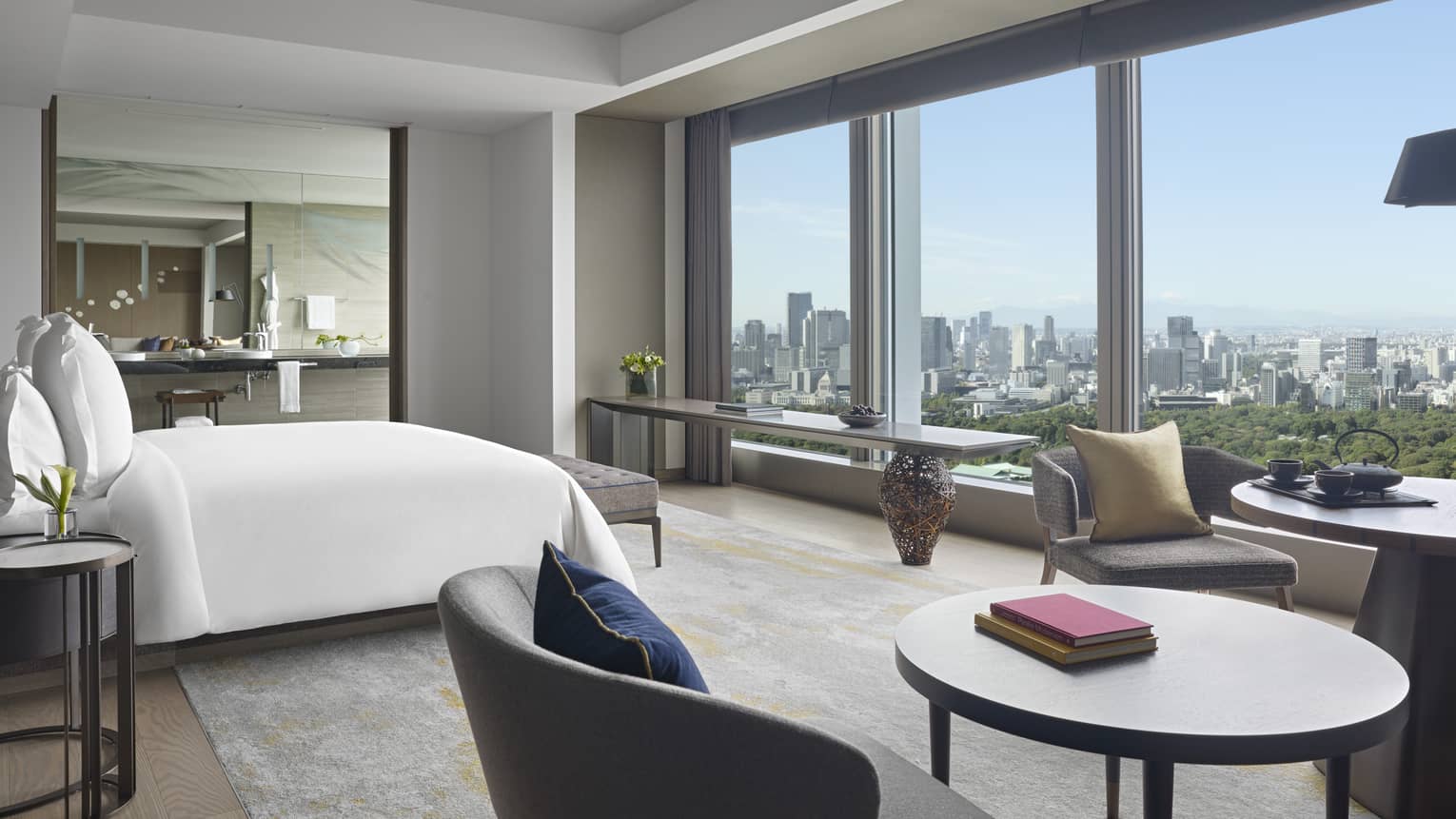 Four Seasons hotel suite with king bed, seating area with round tables and armchairs, side table, large mirror and windows with city views