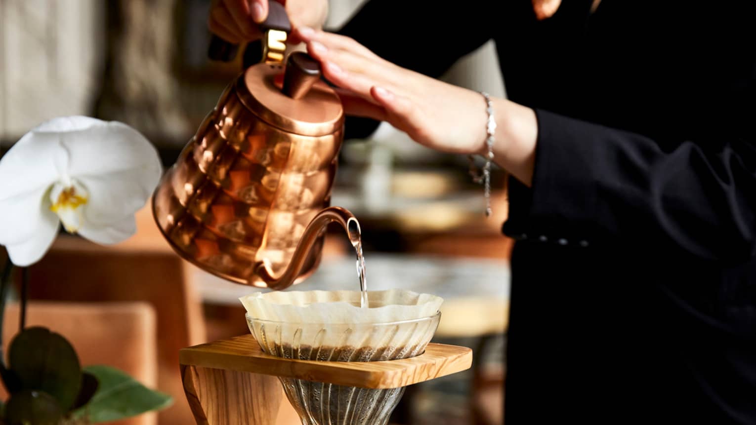 A woman pours water into a filter filled with coffee grounds, which drips into a glass container of hot coffee