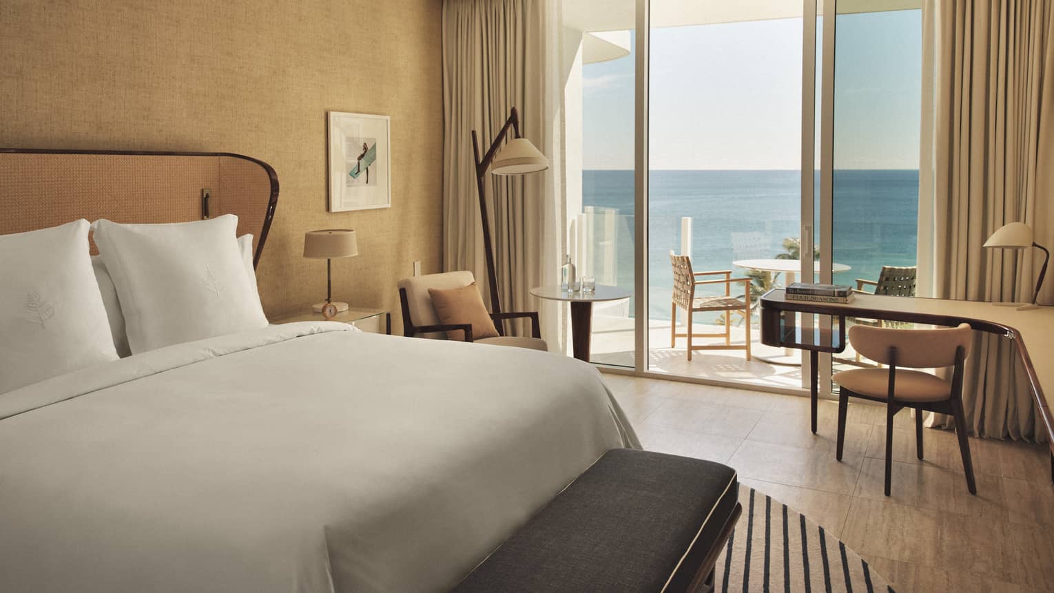 A hotel room with sliding glass door, bed with white sheets, long curved desk, two lamps, two chairs and outdoor seating.