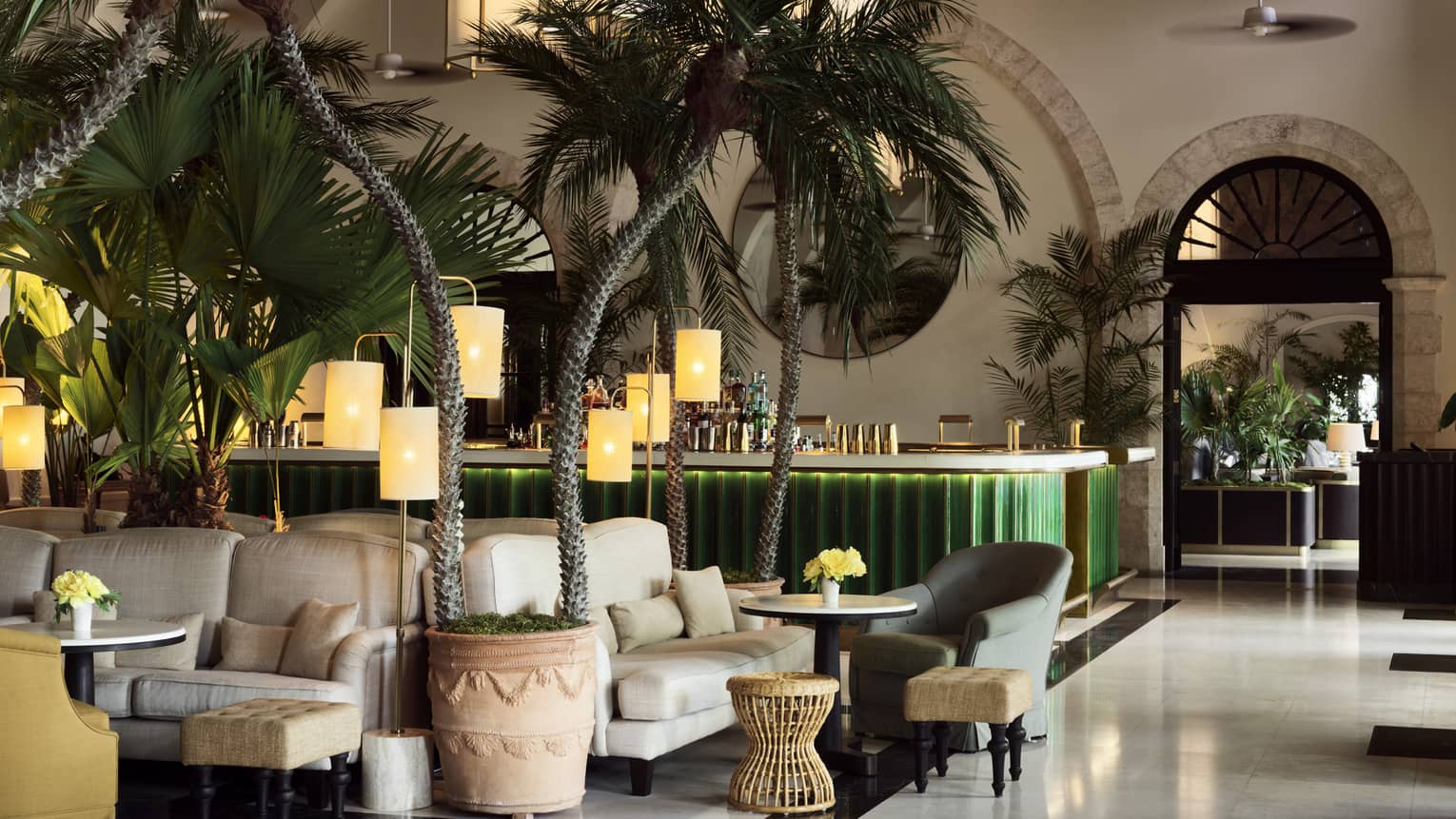 Indoor lounge with palm trees, white upholstered arm chairs, green bar in the background