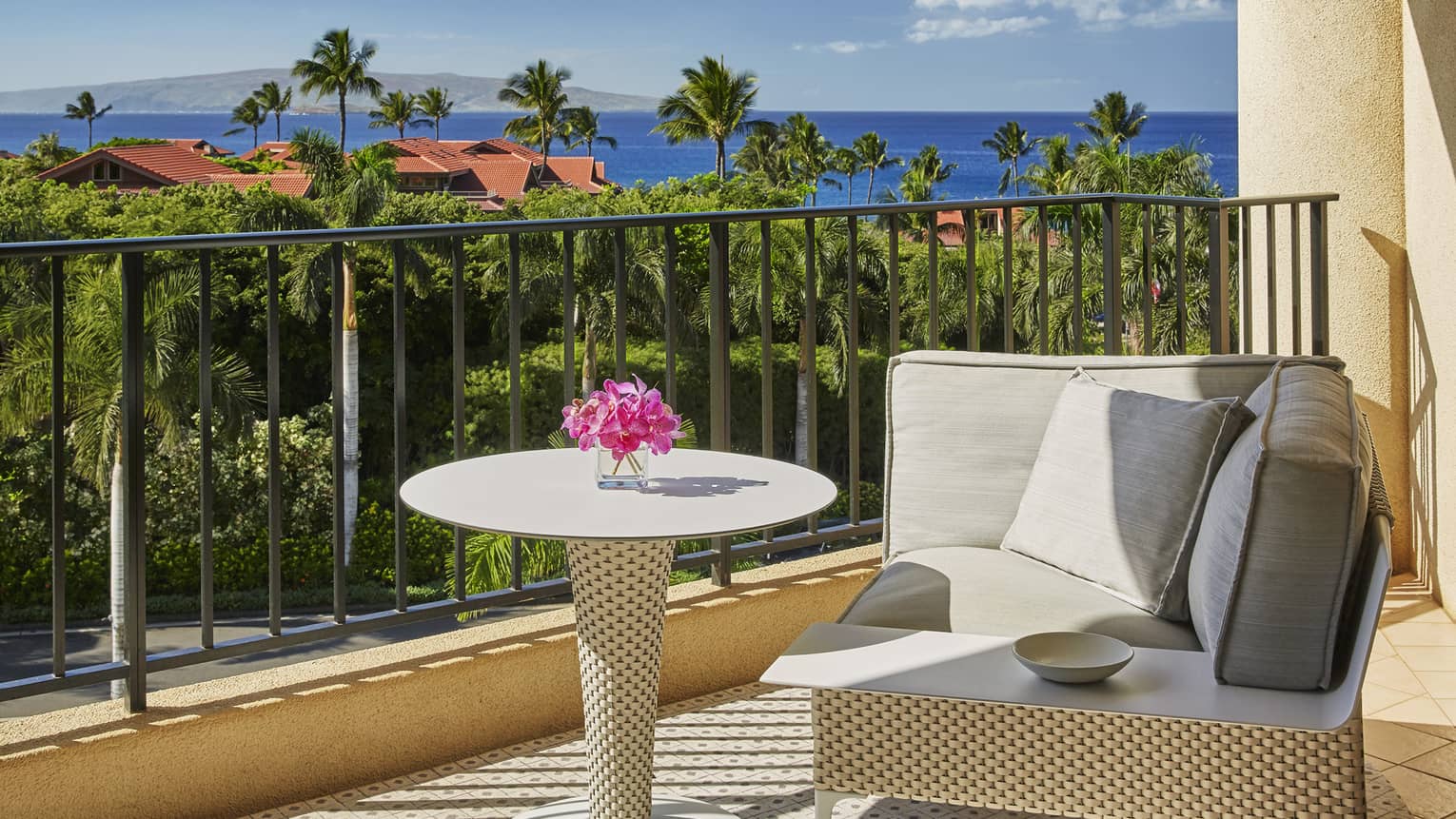 Balcony with cushioned seating, round table and ocean view over lush greenery and rooftops with palm trees