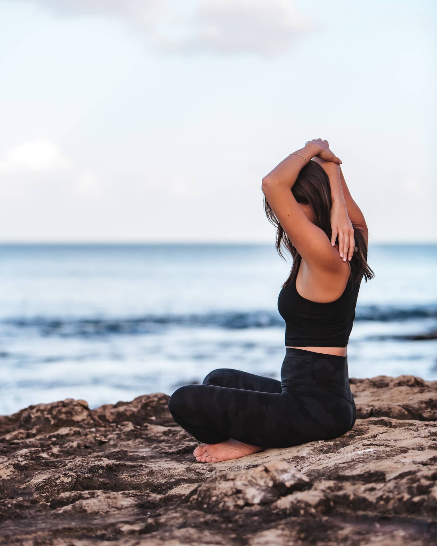 A woman does yoga while sitting on a rock by the ocean