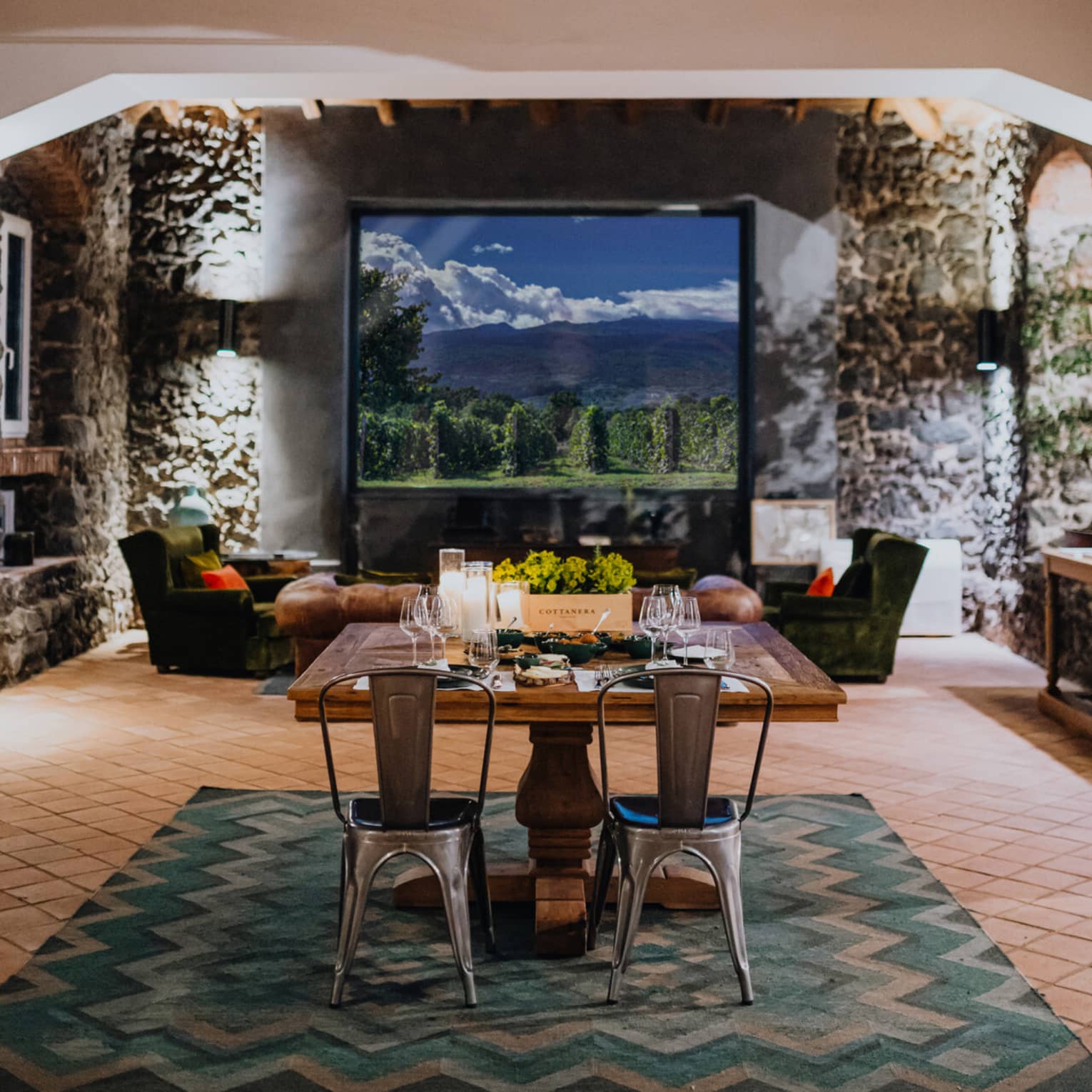 A wooden dining table set with floral arrangements and glassware,  in a room with a window overlooking mountains and a vineyard