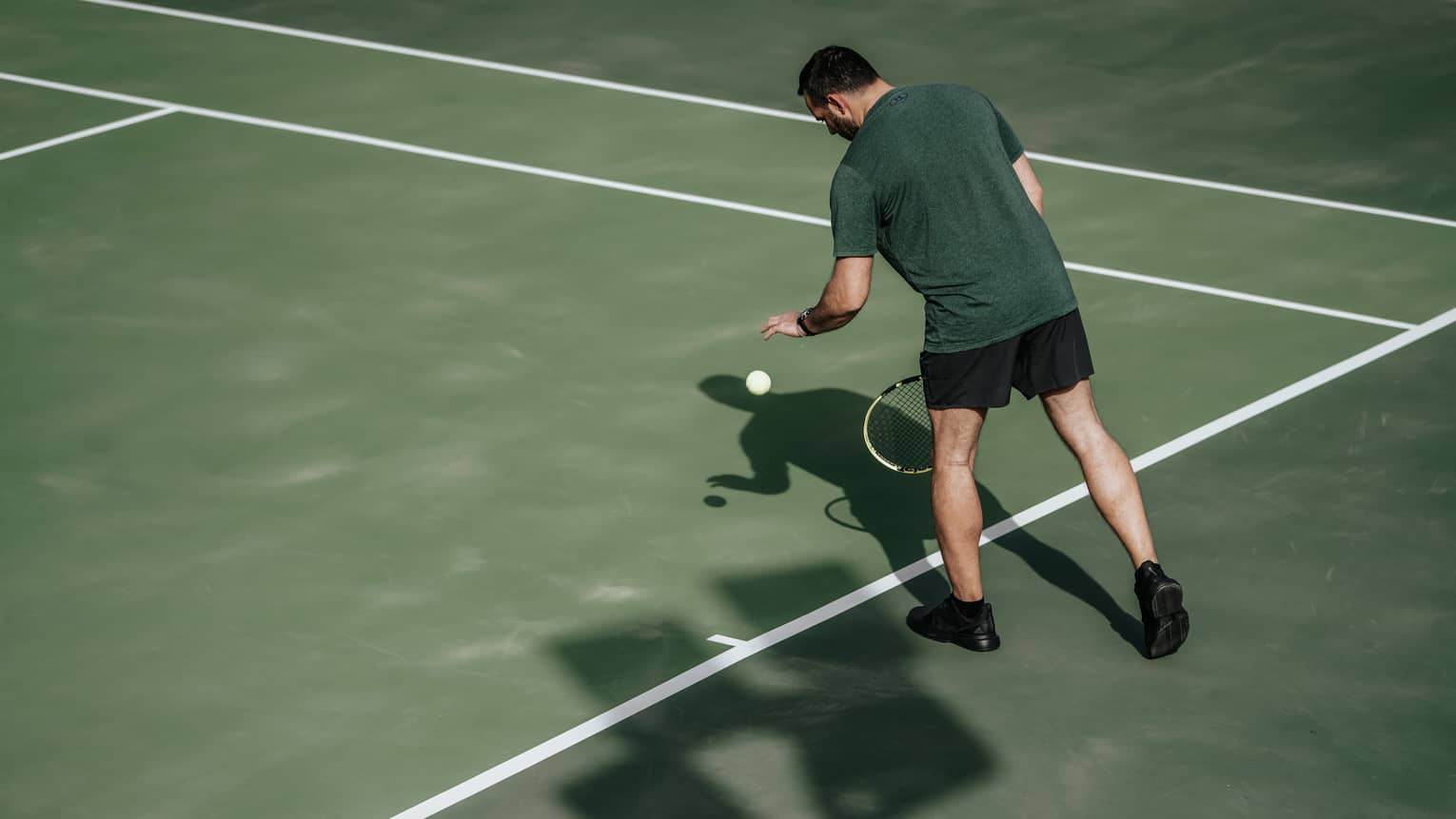 Man on tennis court bouncing tennis ball with left hand, preparing to serve
