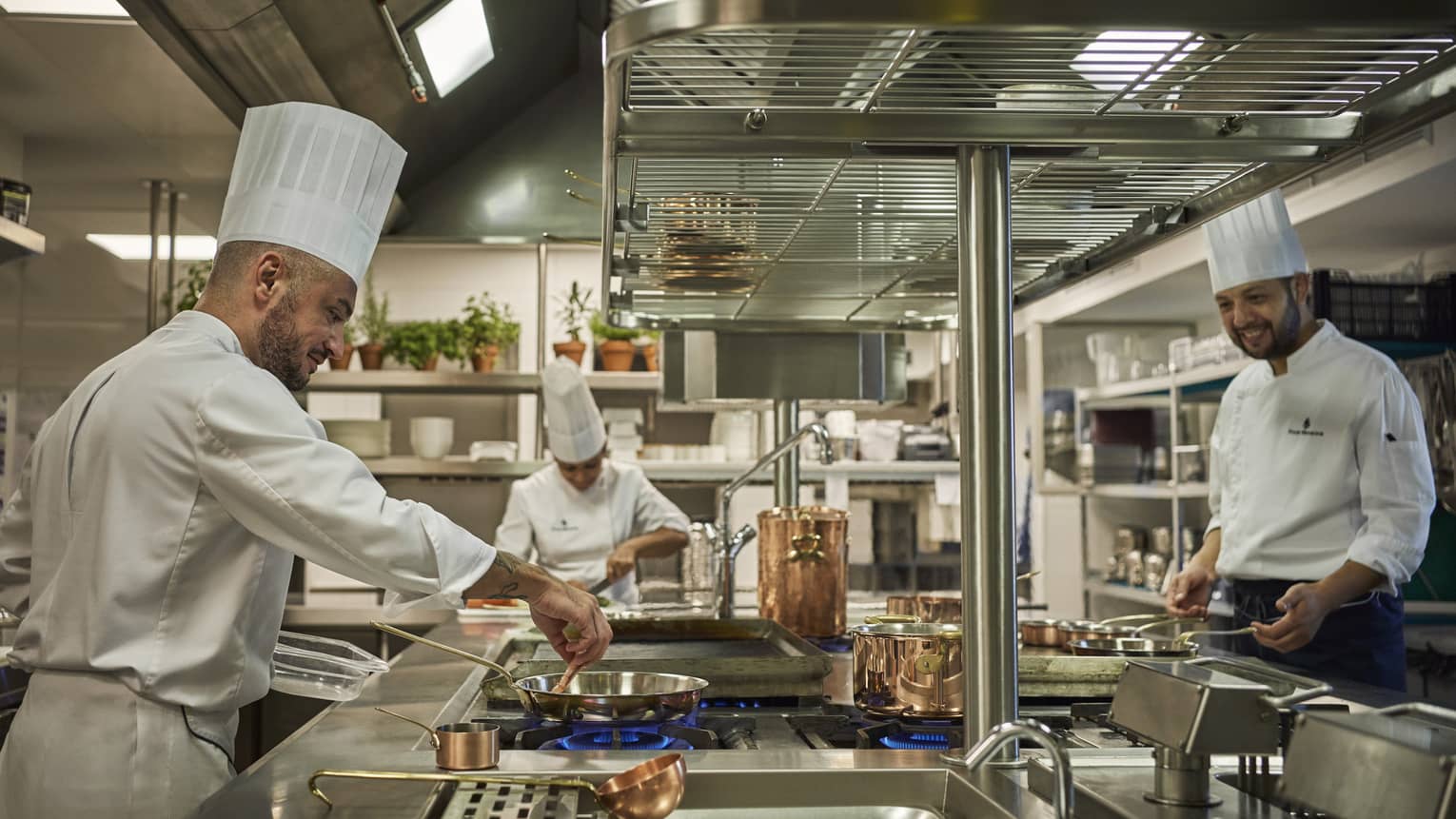 Three chefs with white coats and chefs hats cook in a kitchen