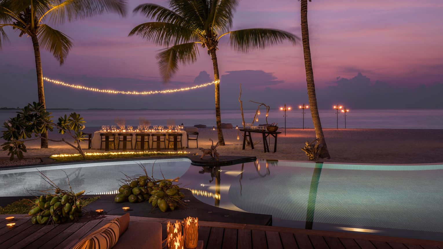 String of lights between palm tree over private dining table on beach by swimming pool at sunset