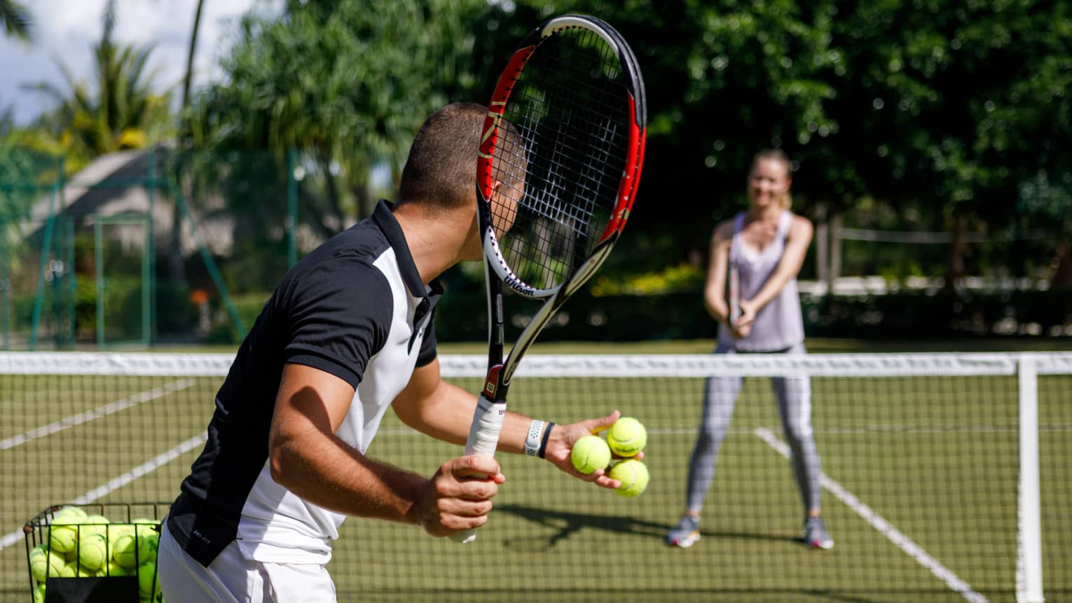 Couple playing tennis on court, man prepares to serve tennis balls to woman ready to swing