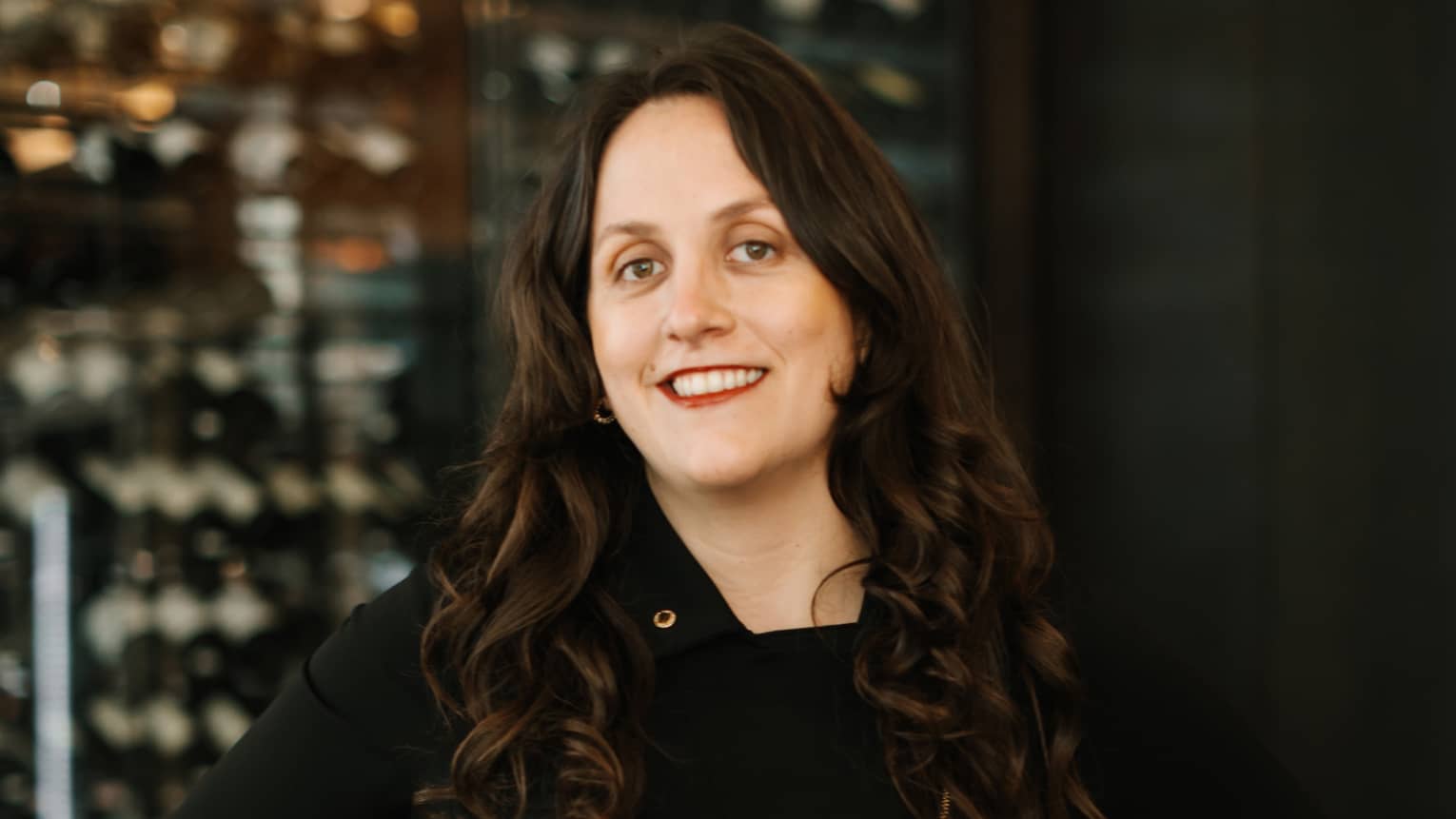 A woman in a black dress in front of shelves of wine.