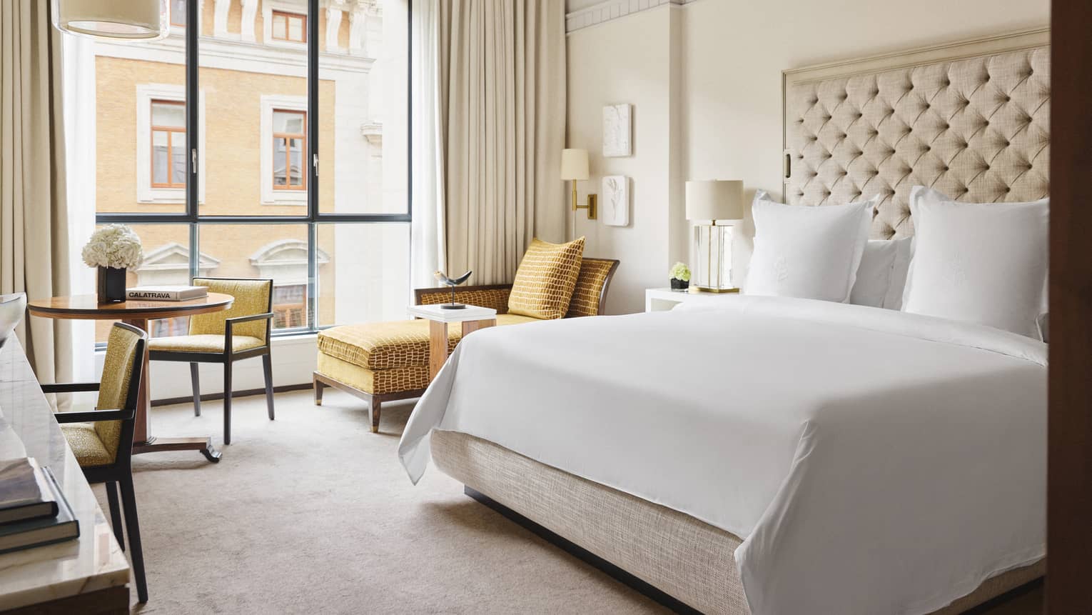 Hotel bedroom with a king bed, tufted headboard, round table, yellow chairs and chaise lounge, horn sculpture and windows overlooking a neighbouring building
