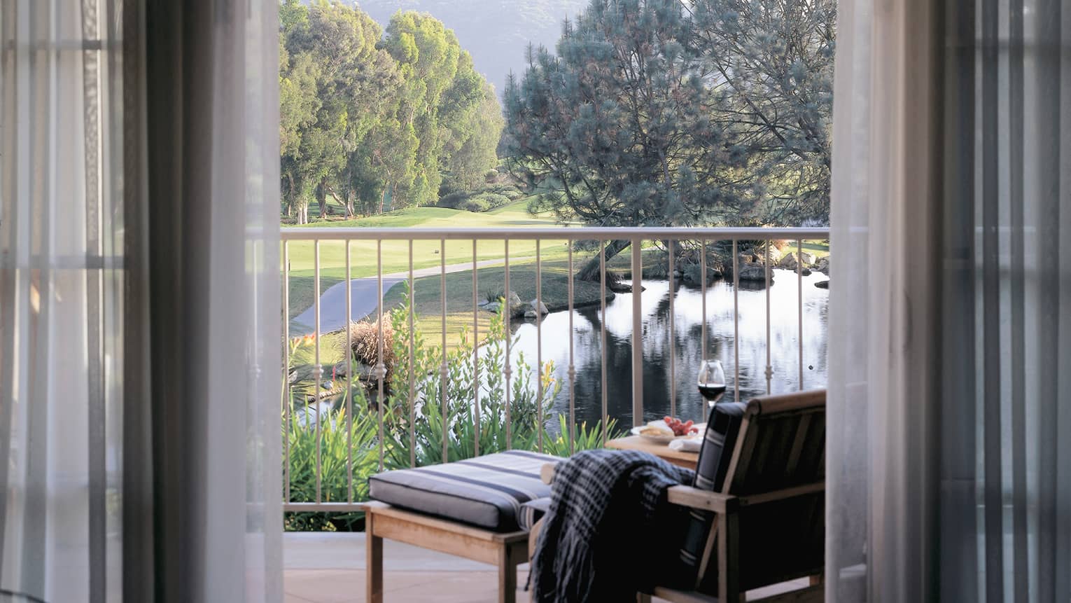 Patio chair with blanket on arm and wine glass on table, looking past railing to pond, garden, trees