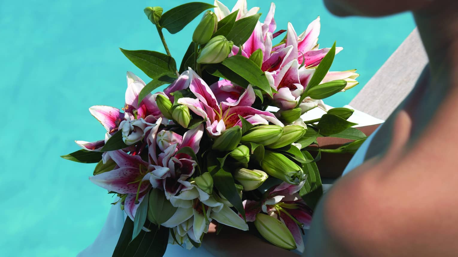Bride holding bouquet of tropical flowers, sits beside sandals on dock over blue lagoon
