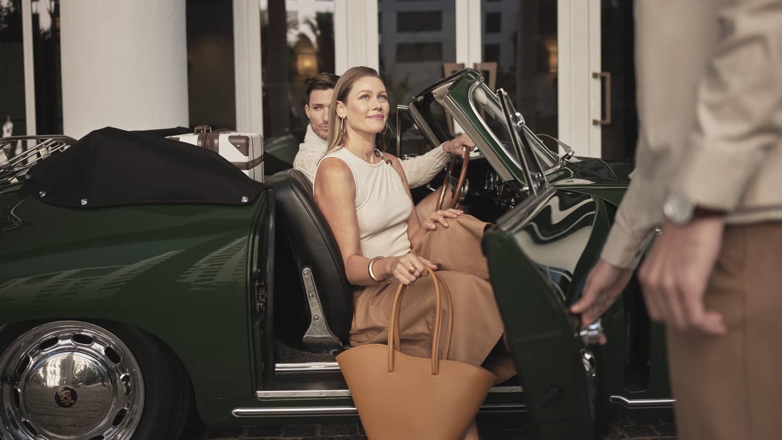A man and woman getting out of a dark green car holding bags, two hotel team members are assisting them.