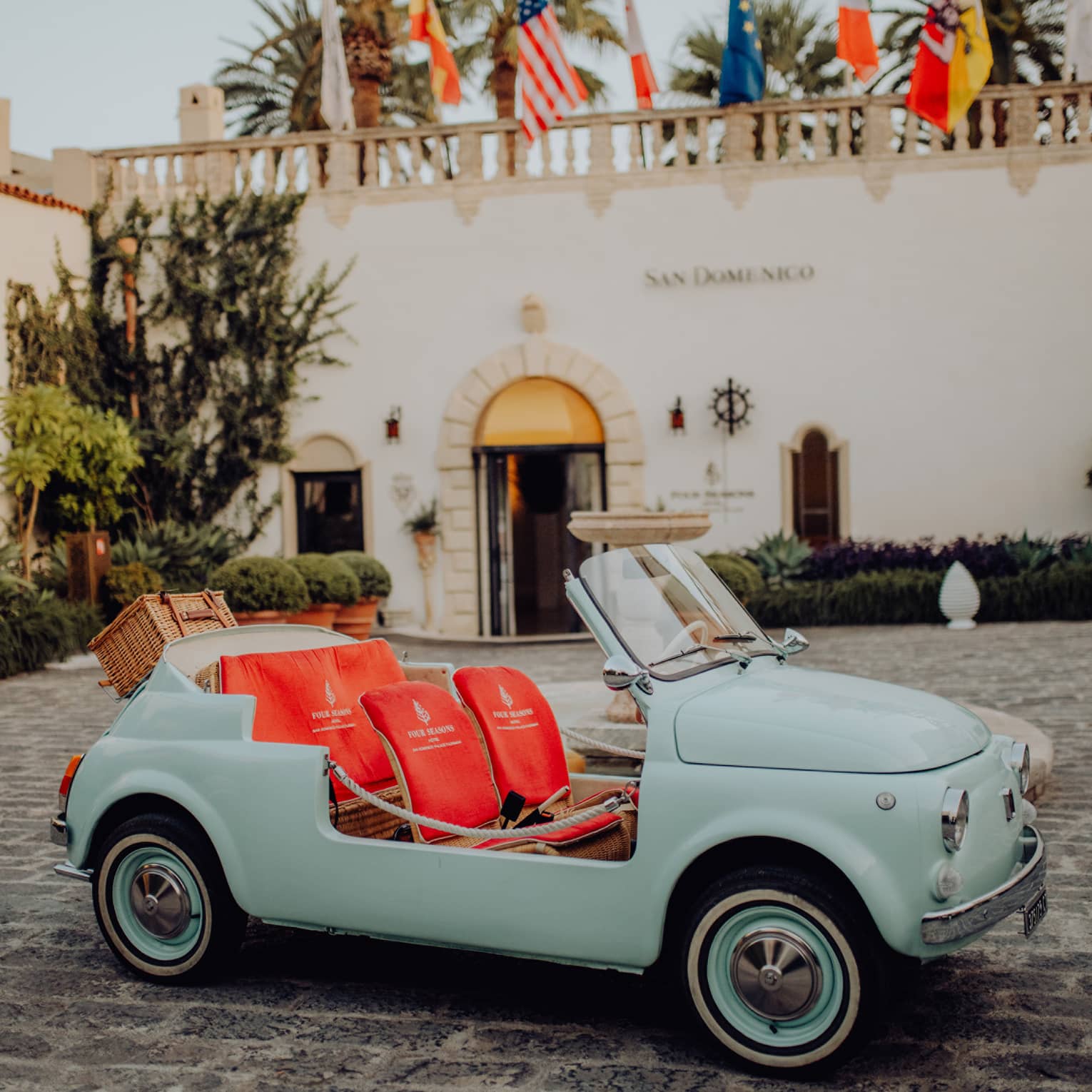 Vintage teal convertible Fiat 500 with bright coral seats parked on a cobblestone driveway in front of San Domenico Palace.  