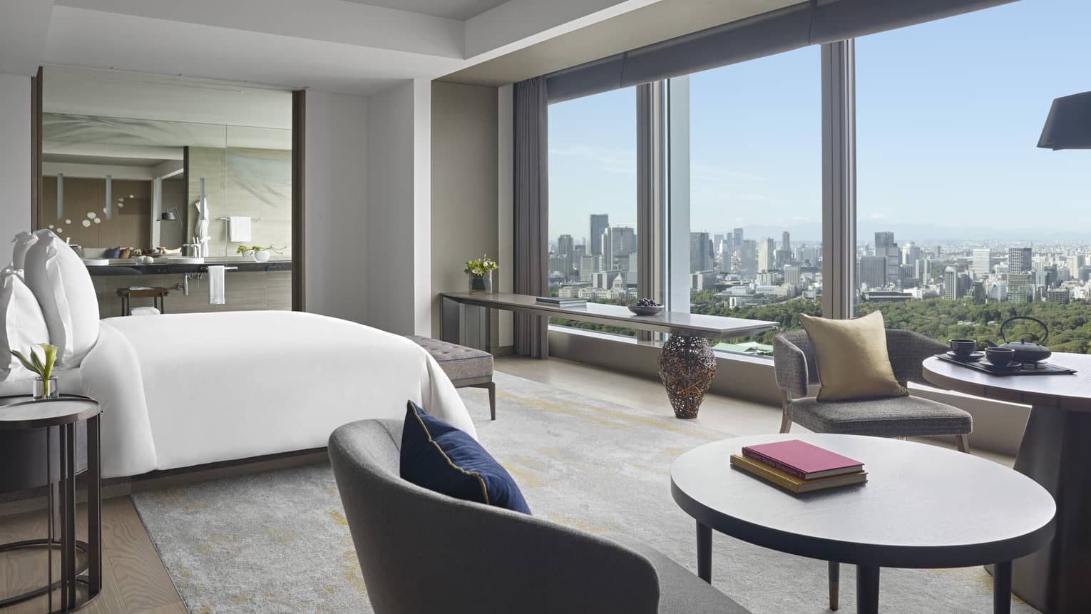 Four Seasons hotel suite with king bed, seating area with round tables and armchairs, side table, large mirror and windows with city views