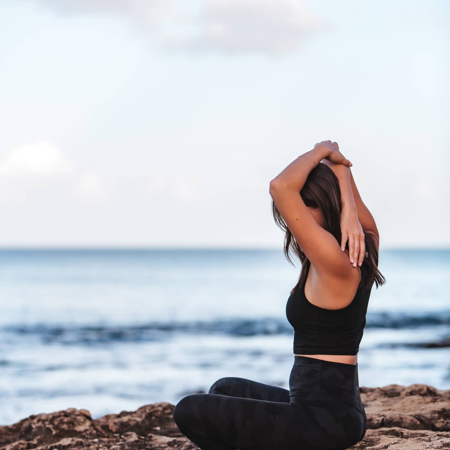 A woman does yoga while sitting on a rock by the ocean