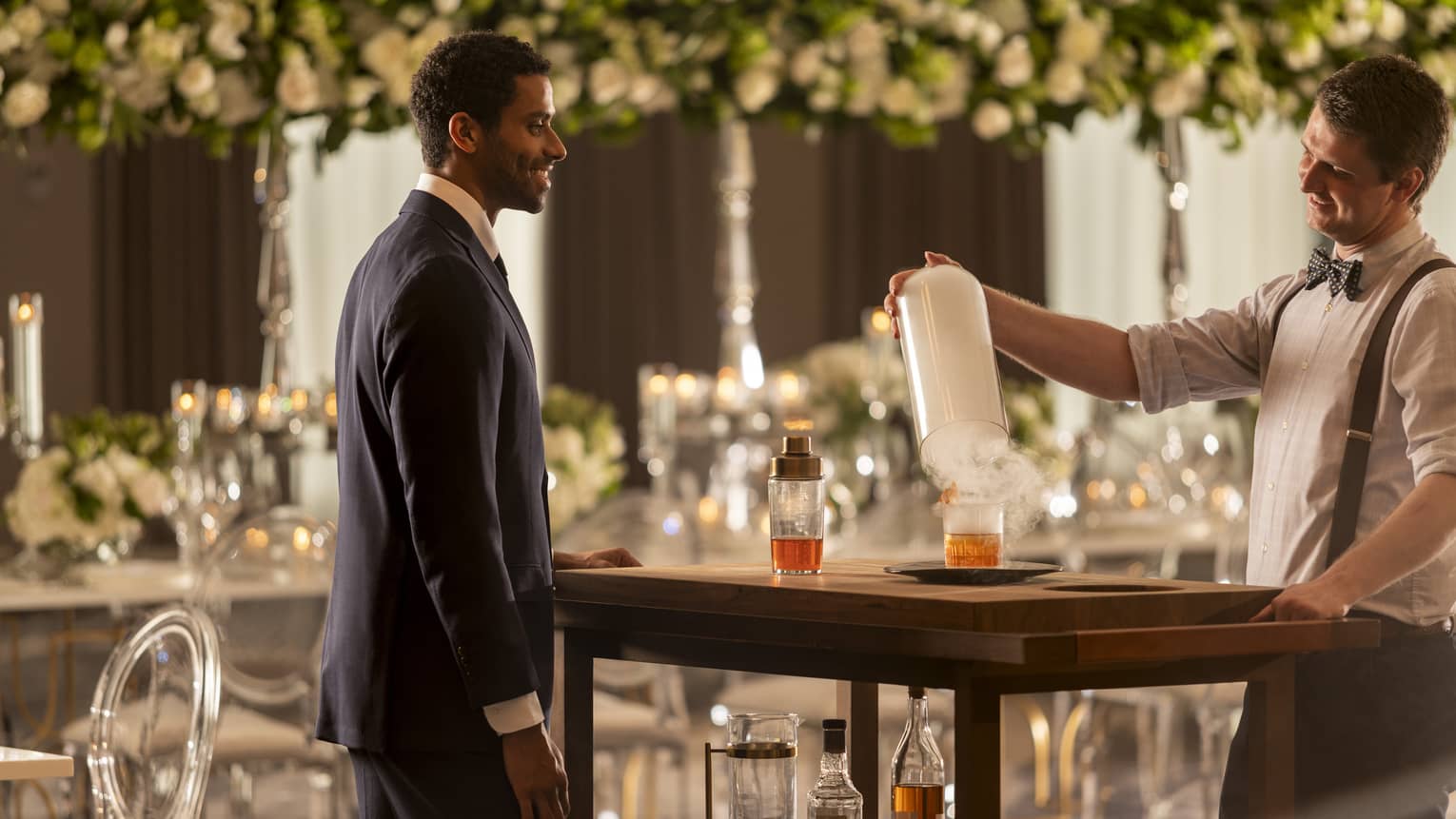 A groom talking to a bartender while the bartender makes a drink, there are flowers hanging in the background.