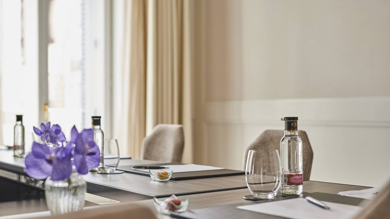 A meeting room with beige walls and curtains, a large window, chairs, and brown tables set with paper, pens, drinking glasses, clear bottles and tiny bowls, plus a vase of purple flowers
