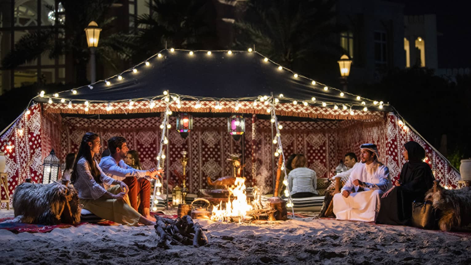 People socialize around fire under Bedouin Tent with lights on sand beach at night