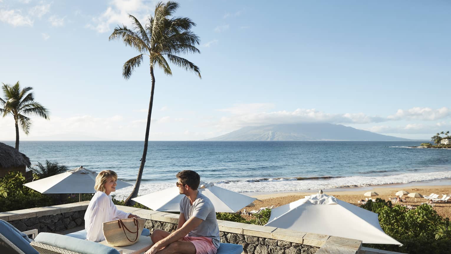 Man in shorts and smiling woman in beach shirt sit on edge of lounge chair, beach and ocean in background