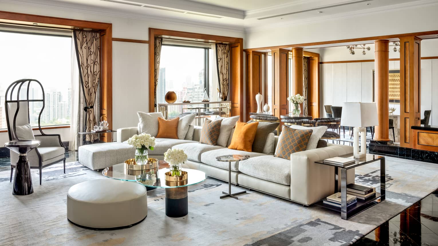 A spacious, luxurious living room with large sectional sofa, orange accent pillows, modern chairs and glass-topped tables. The room features wooden accents and natural light.