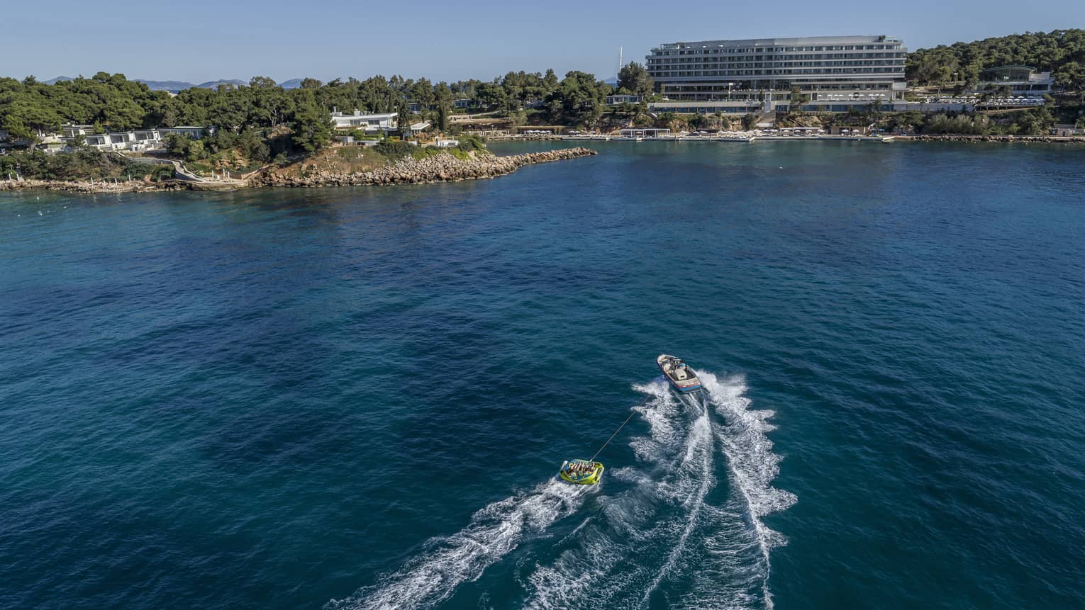 Speedboat pulls raft of people on blue Aegean ocean with building and shoreline in background
