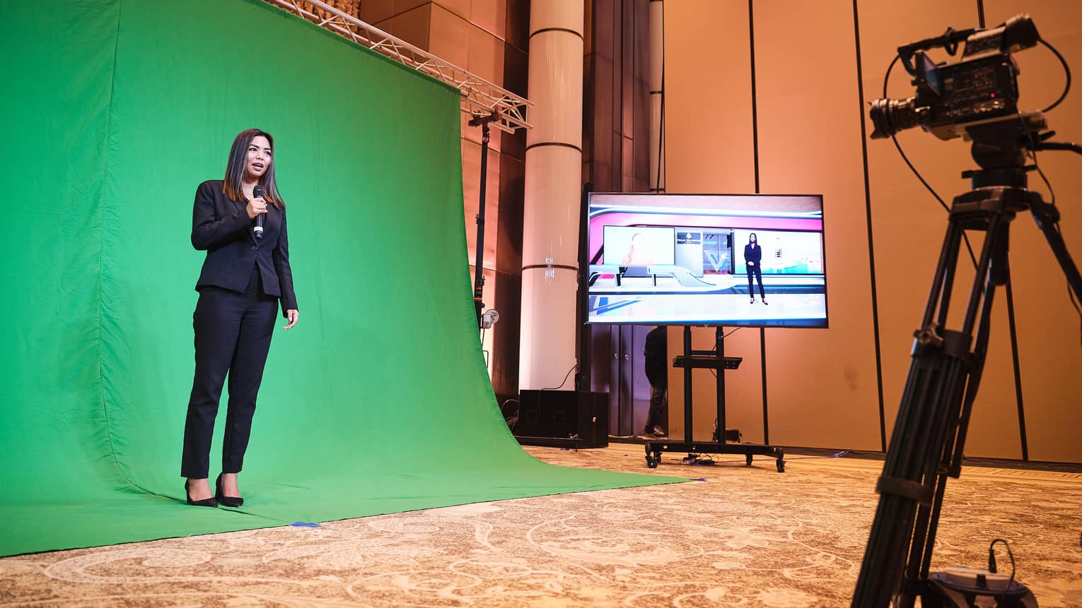 Woman standing and presenting in front of a green screen, with camera on tripod recording her.